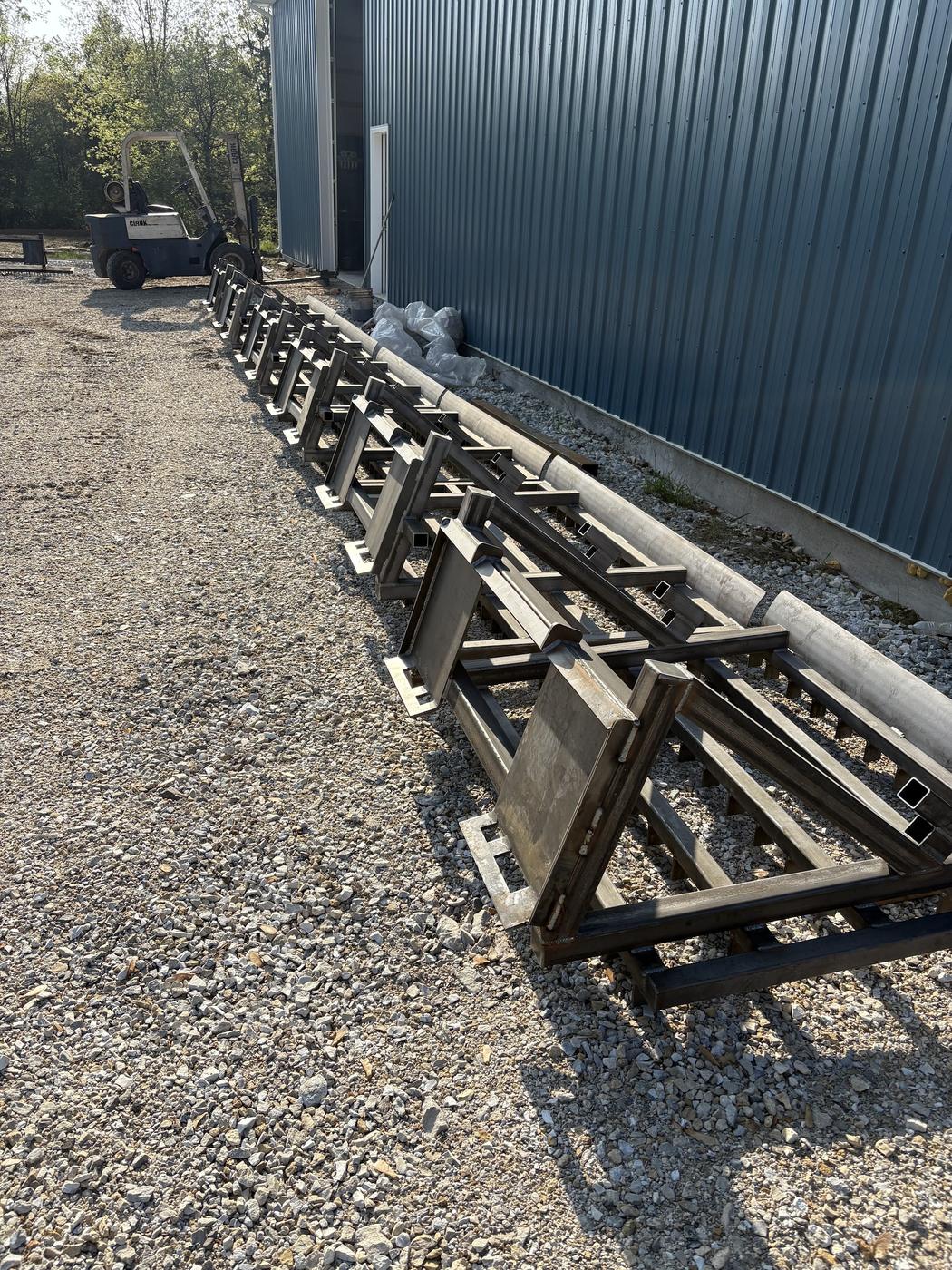 A long row of identical fabricated steel frames laid out on the gravel beside the shop