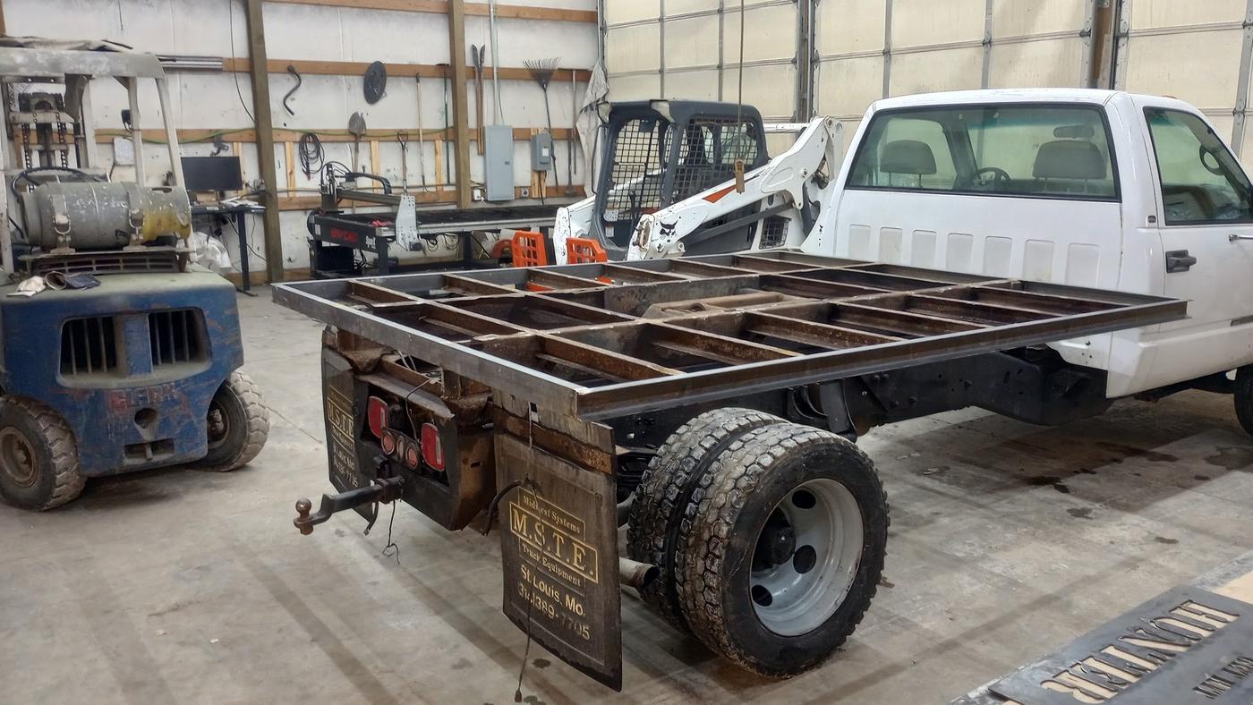 A steel flatbed frame part-built on a white truck chassis inside the shop